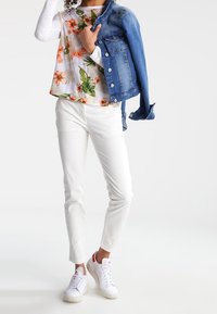 Person in weißen Hosen, floralem Oberteil über einem weißen Langarmshirt, weißen Sneakern, die eine blaue Jeansjacke über einer Schulter hält.