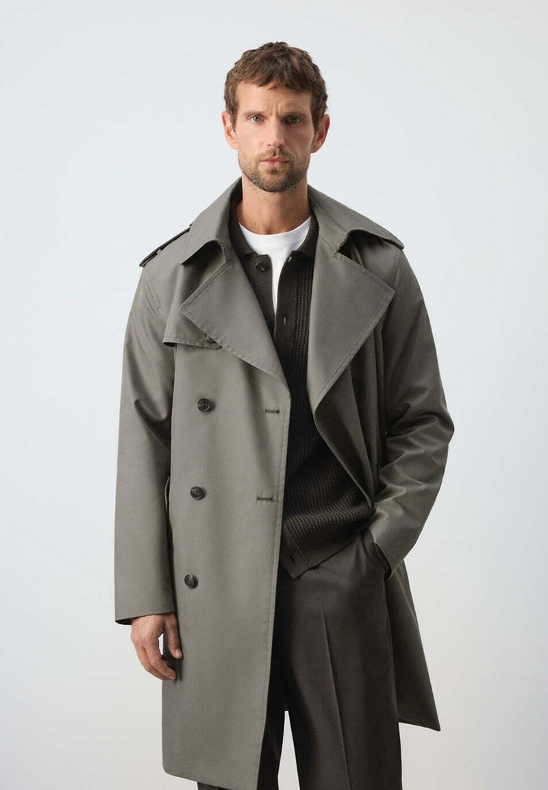 Homme portant un trench-coat gris par-dessus un pull noir et une chemise blanche, debout devant un fond clair uni, une main dans la poche.