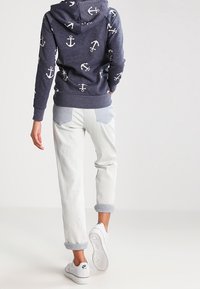 Felpa blu marina con motivo ancorato bianco e dettagli grigi. Jeans chiari con risvolto e sneakers bianche con suola testurizzata, design minimale.