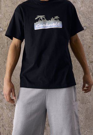 Person trägt ein schwarzes Carhartt T-Shirt mit Palmen- und Stadtsilhouette als Print, kombiniert mit hellgrauen Shorts vor einem Steinmauer-Hintergrund.