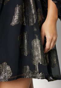 Svart klänning i tyg med metalliskt guldblommönster, med en flytande form och texturerad yta. Hand som håller i tyget vid fållen.