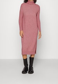 Robe en tricot à manches longues dans un rose chiné avec col montant et fentes sur les côtés, associée à des bottines noires à semelles épaisses.
