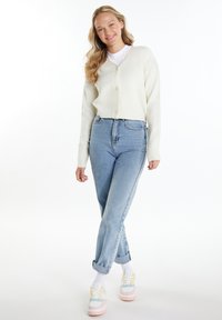 Weißer, gestrickter Cardigan mit V-Ausschnitt und fünf Knöpfen, kombiniert mit hochgeschnittenen hellblauen Jeans und mehrfarbigen Sneakers.