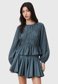 Top peplum e gonna plissettata in blu polveroso. Tessuto morbido e strutturato con dettagli arricciati. Maniche lunghe con polsini elastici e orlo fluente.