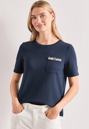 Blondynka w granatowym krótkim topie z dekoracyjną kieszenią i białych spodniach, stojąca z rękami w kieszeniach.