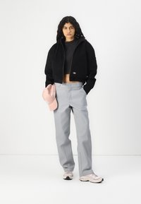 Giacca nera cropped con zip, pantaloni grigi a gamba dritta e cappellino rosa chiaro. Tessuto testurizzato e dettagli del logo discreti; sneaker con accenti chiari.