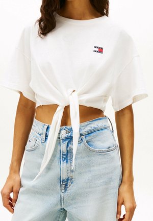 Kobieta w białym krótko przyciętym t-shircie z wiązaniem z przodu z małym logo Tommy Jeans oraz jasnoniebieskich dżinsach z wysokim stanem.