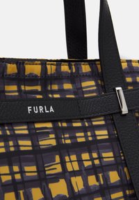 Des poignées en cuir noir sont attachées à un tissu à motifs jaunes et foncés, avec le nom de la marque « FURLA » en blanc sur une sangle noire.