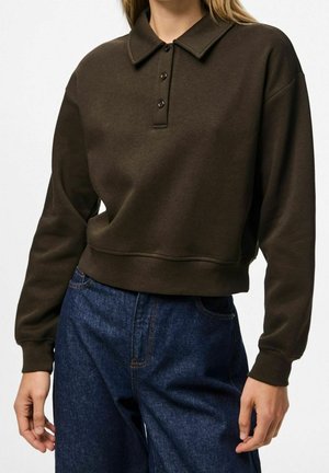 Vrouw die een donkerbruine polo met lange mouwen en een high-waist blauwe spijkerbroek draagt, staand met ontspannen handen langs haar zij.