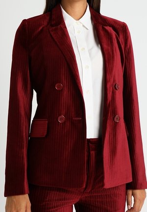 Femme portant un blazer en velours rouge profond à double boutonnage et un pantalon assorti avec une chemise blanche boutonnée en dessous.