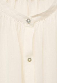 Blouse couleur crème avec un col rond, des plis verticaux et des boutons argentés à l'avant. Fabriquée en tissu léger.