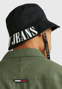 Persona con un sombrero de cubo negro con el texto "JEANS" en el ala y una camisa verde oliva con una pequeña etiqueta de bandera rojo, blanca y azul en la parte de atrás.