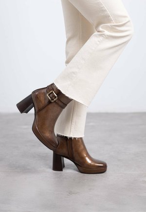 Botas de cuero marrón hasta el tobillo con un acabado brillante, un tacón grueso y un detalle de hebilla. Combinadas con pantalones acampanados de color blanco roto.