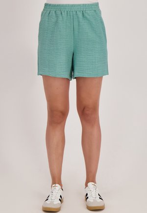 Persoon in teal elastische taillebroek en witte sneakers met zwarte strepen, staande tegen een effen lichte achtergrond.