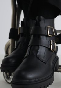 Black leather ankle boots with two gold buckles, textured finish, chunky soles, and a rugged design, positioned near a wheelchair.