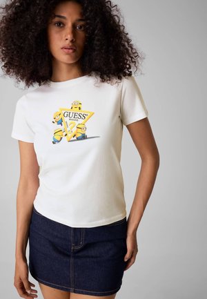 Donna con capelli ricci che indossa una t-shirt bianca con il logo giallo dei Minions e Guess e una gonna di jeans blu scuro.