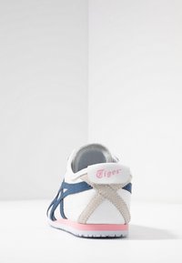 Vit sneaker med blå och beige detaljer, med en rosa logotyp på hälen och en strukturerad yttersula. Låg profil och snörningsdesign.