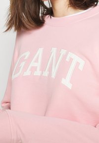 Sweatshirt rose clair en tissu doux avec des lettres blanches brodées formant "GANT" sur la poitrine. Poignets et col en tissu côtelé.