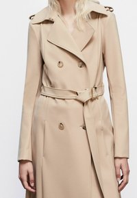 Gabardina beige confeccionada en tela suave, con diseño de doble botonadura, botones prominentes y cinturón, con hombreras en los hombros.