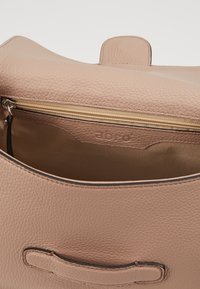 Růžová kožená kabelka s texturovaným povrchem, doplněná zlatým zipem a vnitřní kapsou. Logo vyražené uvnitř. Minimalistický design.