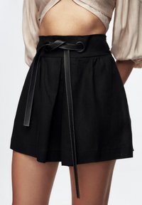 Sort kort nederdel lavet af et tekstureret stof, med bred taljebånd, plisseringer og et sort bindebånd med kontrasterende syningsdetaljer.