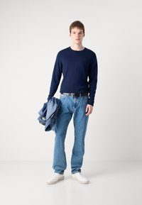 Cămașă navală cu mânecă lungă, blugi din denim de culoare albastru deschis, adidași albi, curea neagră, ținând o jachetă din denim. Design simplu, casual.