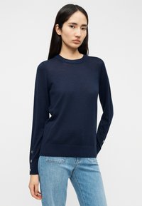 Μπλε πλεκτό πουλόβερ με στρογγυλή λαιμόκοψη, μακριά μανίκια και κουμπιά στα μανίκια. Μαλακή υφή και άνετη εφαρμογή, συνδυασμένο με ανοιχτό μπλε τζιν.