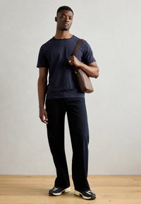 Marineblaues T-Shirt, dunkle Jeans und mehrfarbige Sneakers. Dunkelbraune Umhängetasche mit geometrischem Design. Holzboden mit neutralem Hintergrund.