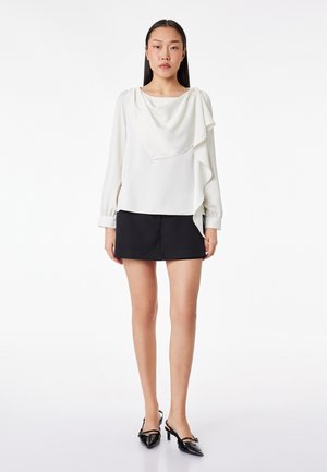 Kobieta ubrana w białą bluzkę z długim rękawem z detalem drapowanym oraz czarną mini spódnicę, w zestawieniu z czarnymi szpilkami wsuwanymi, stojąca na białym tle.