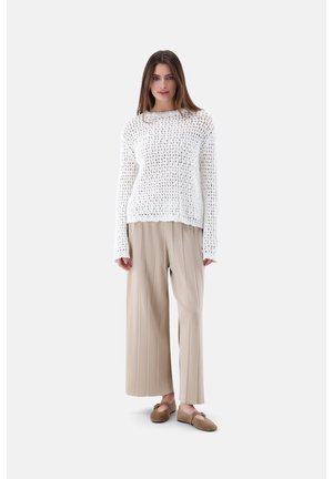 Femme portant un pull en maille blanche à mailles ouvertes, un pantalon plissé beige à jambes larges et des chaussures marron sans lacets, debout devant un fond uni.