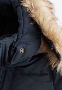 Chaqueta acolchada azul marino con capucha de piel sintética desmontable. Presenta botones a presión plateados y forro suave negro. Acento de piel texturizada a lo largo de la capucha.