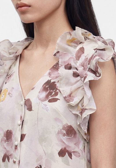 Γυναίκα που φοράει αμάνικη φλοράλ μπλούζα με βολάν στους ώμους και κουμπιά καλυμμένα με ύφασμα, δείχνοντας τον λαιμό, τον ώμο και το επάνω μέρος του στήθους.