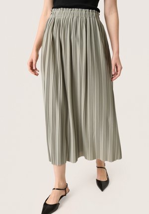 Femme portant une jupe midi plissée gris clair taille haute et des ballerines noires à bout pointu avec bride à la cheville, debout sur un fond clair.