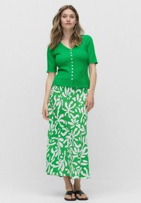 Green textured cardigan with short sleeves, paired with a long green skirt featuring a white floral pattern. Black sandals complete the outfit.