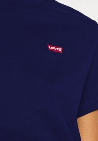 Námořnická modrá bavlněná tričko s krátkým rukávem a kulatým výstřihem, se červenou štítkem loga Levi's na hrudi. Hladká textura a jednoduchý design.