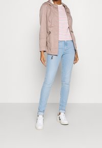 Woman wearing light pink jacket, white and pink striped shirt, light blue jeans, and white sneakers standing against a plain background.