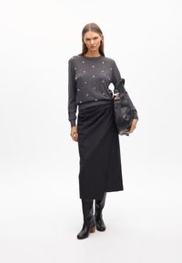 Grauer gestrickter Pullover mit runden Verzierungen, kombiniert mit einem schwarzen Wickelrock und kniehohen schwarzen Lederstiefeln. Schwarze Tasche mit geflochtenem Trageriemen.