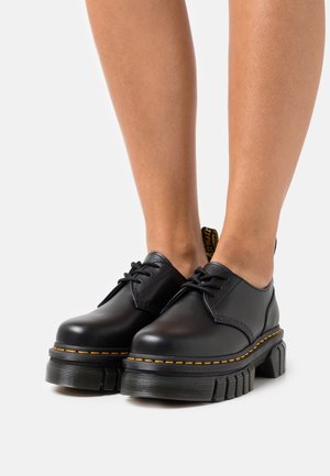 Jambes portant des chaussures en cuir noir à lacets avec des semelles épaisses à rainures et des détails de couture jaune sur un fond blanc.