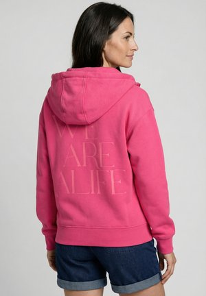 Frau mit dunklen Haaren, die ein leuchtend pinkes Kapuzensweatshirt mit dem Text "WE ARE A LIFE" auf dem Rücken und dunkelblaue, hochgekrempelte Jeansshorts trägt.