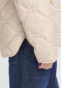 Veste matelassée beige avec un motif ondulé et un ourlet arrondi, associée à un denim foncé. Dispose d'un éclat doux et de détails de couture visibles.