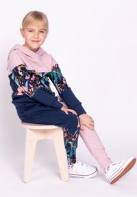 Wielokolorowy dres z różowym kapturem i rękawami w kwiatowy wzór, w połączeniu z granatowymi spodniami. Ubrany z białymi trampkami, siedzący na stołku.