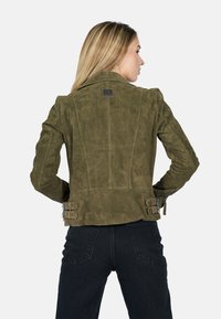 Olijfgroene suede jas met een getailleerde pasvorm, voorzien van gespen op de taille en een spitse kraag. De textuurafwerking versterkt het totale ontwerp.