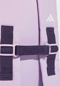 Sac à dos violet en tissu texturé, avec des sangles et des boucles violet foncé. Logo blanc sur le côté. Design compact et fonctionnel.