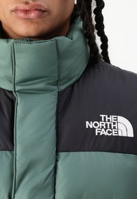 Pufasta jakna u tamnozelenoj i crnoj boji, s visokim ovratnikom, kviltanom teksturom i bijelim logom North Face na prsima.