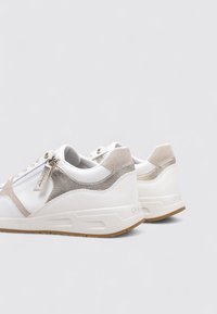 Zapatillas blancas con un acabado liso, que cuentan con detalles en beige y elementos metálicos. Forma redondeada y suela de goma texturizada. Cierre con cremallera.