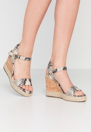 Sandalias de cuña de corcho trenzado con tiras de piel de serpiente en blanco y negro, hebilla en el tobillo y suela de cuerda estilo esparto en los pies de una persona.