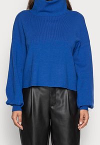 Pull bleu court avec un col roulé côtelé haut, des manches larges et une texture douce, associé à un pantalon en cuir noir.