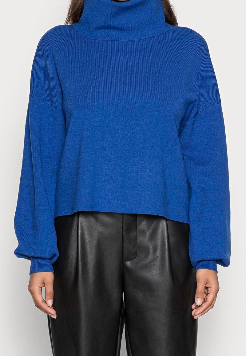 Pull bleu court avec un col roulé côtelé haut, des manches larges et une texture douce, associé à un pantalon en cuir noir.