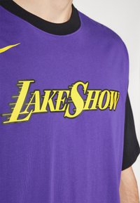 Tricou scurt cu mânecă violetă, având inscripția bold "LAKE SHOW" în galben. Gulerul negru contrastează cu textura netedă a materialului.