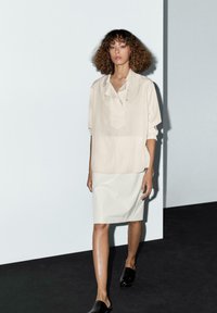 Beige, losse blouse met een knoopsluiting, gecombineerd met een witte knie-lange rok en zwarte instappers, geplaatst tegen een minimalist background.
